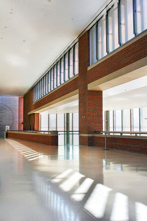 Sunlit interior corridor with brick walls and tall windows with blinds, casting striped shadows across a polished floor.